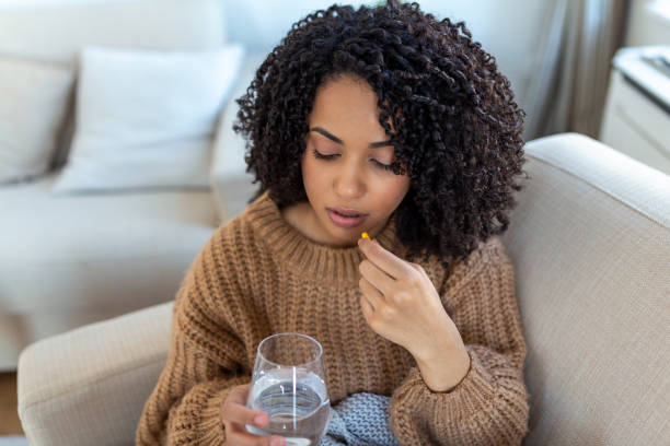 concept de souffrir d’une douleur terrible. belle, triste, bouleversée, malheureuse, troublée, femme faible aux cheveux noirs, portant des vêtements décontractés est assise sur un canapé et prend une pilule - painkiller photos et images de collection