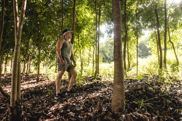 un homme asiatique en forme prend une pause et s’appuie sur un arbre au milieu d’une forêt pendant une course de trail. concepts cardio et aventure. - sportsman tree people recreational pursuit photos et images de collection