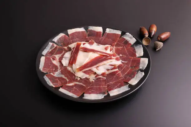Ration of 100% Dehesa de Extremadura acorn-fed Iberian ham on a black plate and black background decorated with acorns