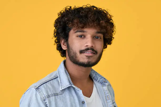 Young curly smart indian cool guy standing isolated on yellow background. Ethnic cool handsome man wearing denim shirt looking at camera posing for headshot close up face portrait.