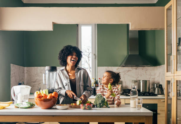 An African-American Single Mother Preparing Vegan Lunch In The Kitchen And Smiling With Her Little Daughter stock photo