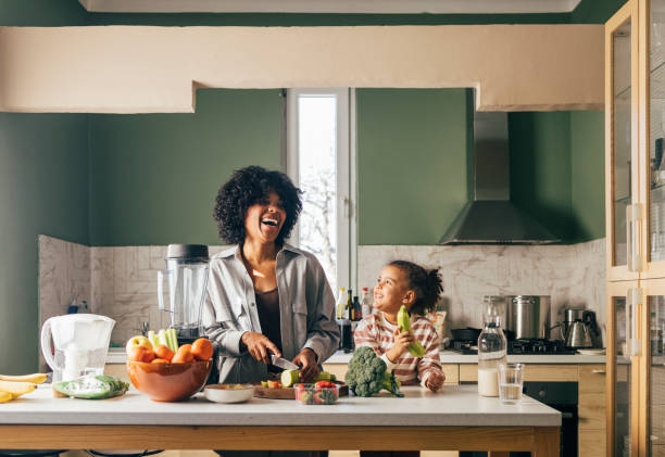 uma mãe solteira afro-americana preparando almoço vegano na cozinha e sorrindo com sua filhinha - child mixed race person little girls human gender - fotografias e filmes do acervo