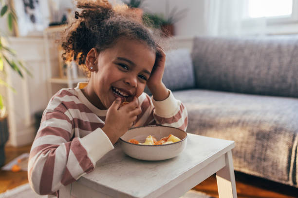 un ritratto di una simpatica ragazza afro-americana seduta nel suo salotto sorridente e godendo di mangiare un'arancia - little girls small eating breakfast foto e immagini stock