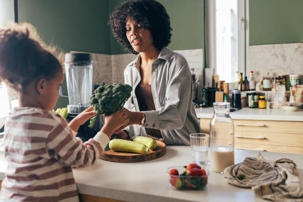 una madre afro-americana che prepara il pranzo con sua figlia usando solo verdure crude biologiche - blender concepts red black foto e immagini stock