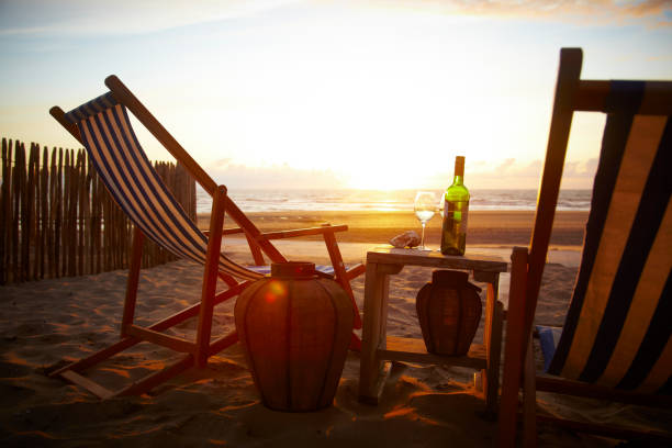 The setting sun is reflected in the water of the sea. There are chairs and a small table with wine glasses and a bottle on it. The strand is empty. On the left you can see a small fence.