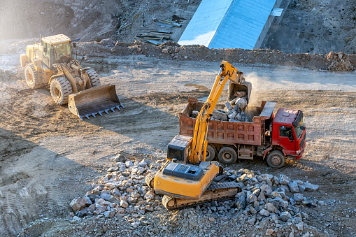 Yellow color excavator and bulldozer are loading some rocks to red color earth dump truck in the construction site.