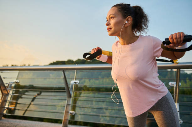 A sporty woman, female athlete exercising with suspension straps outdoor, doing exercises on arms and shoulders muscles while practicing body weight training on the city bridge early in the morning mujer deportista, atleta femenina que hace ejercicio con correas de suspensión al aire libre, haciendo ejercicios en los músculos de los brazos y los hombros mientras practica el entrenamiento con peso corporal en el puente de la ciudad temprano en la ma