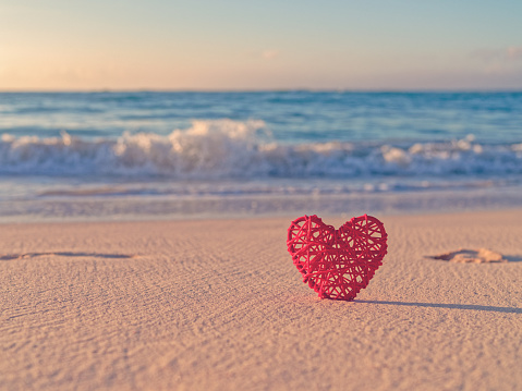 The figurine of the heart is in the sand on a luxurious sandy beach. Footprints in the sand. Waves in the sea. Dawn in the sky. Tropical layout for Valentine's Day.