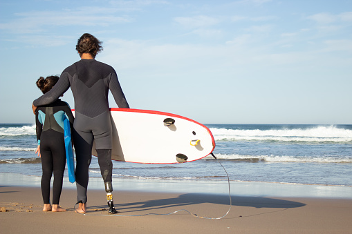 Loving father with mechanical leg with daughter on beach. Back view of mid adult man and little dark-haired girl carrying surfboards, looking at water. Family, leisure, active lifestyle concept