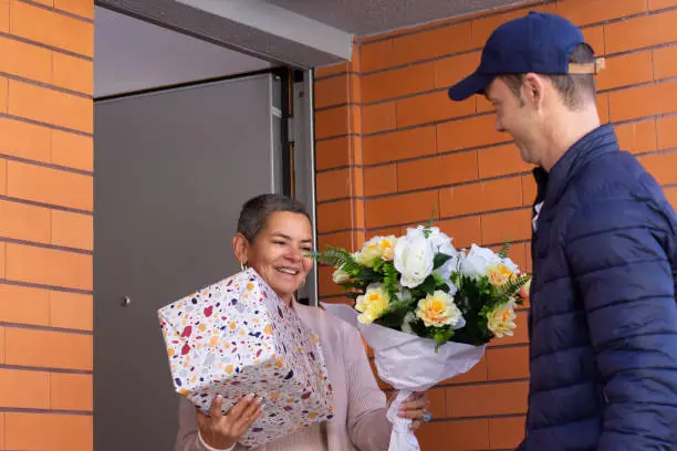 Smiling deliveryman giving bouquet and gift to female customer. Mid adult man in cap and dark blue coat delivering flowers on sunny day. Work, service, shipment concept
