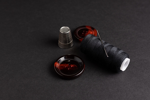 Sewing kit showing burgundy buttons, black thread, needle and thimble on black background.