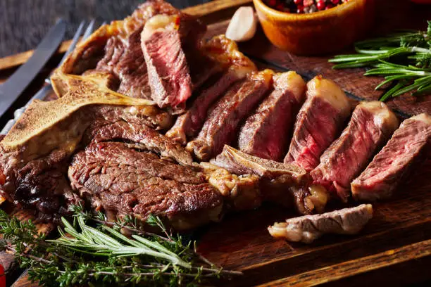 fried sliced porterhouse steak, t-bone steak on a rustic wooden board with tomatoes, herbs, peppercorn, cutlery on a dark wooden table, horizontal view from above, close-up