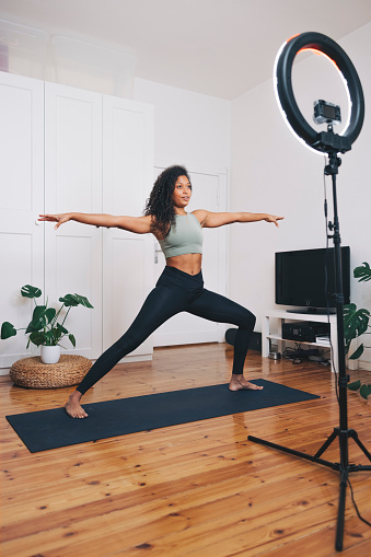 Woman conducting online exercise class using mobile phone with ring light. Fitness instructor live streaming yoga workout from home.