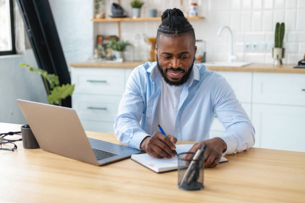 istock Smiling African American man uses laptop computer for online learning remotely at home taking notes on notepad during online lesson 1367719831