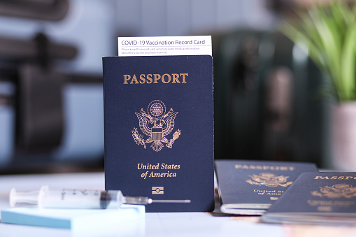 Close-up view of a USA passport with COVID-19 vaccination record card and syringe. Luggage defocused in background. Concepts.