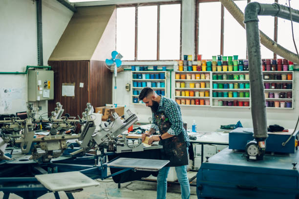 männlicher arbeiter, der tinte auf den rahmen drückt, während er die druckmaschine in einer werkstatt benutzt - silk screen fotos stock-fotos und bilder