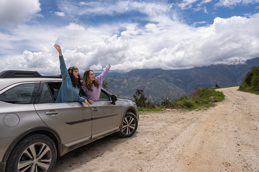Two young women protruding from a car window with the arms raised gesturing freedom in the mountains