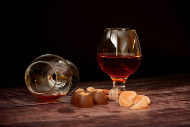 dos vasos de coñac, canela, chocolates y rodajas de mandarina sobre una mesa de madera. - liqueur glass fotografías e imágenes de stock