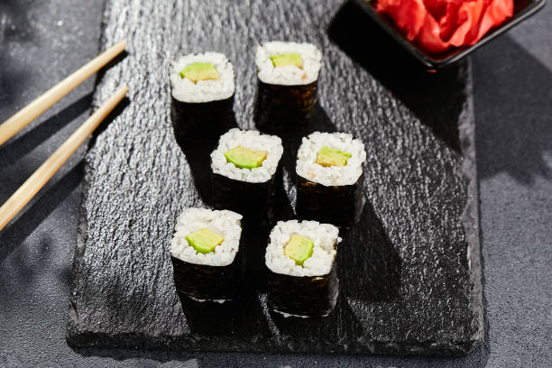 Avocado hosomaki on dark stone table. Hosomaki with avocado. Sushi roll with avocado inside. Style concept japanese menu with black background, leaves and hard shadow.