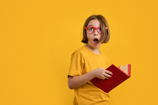 Shocked Child School Girl with Book with Wide Open Mouth and Look to the Side. Little Girl 8-10 Years Old in Yellow T-shirt and Red Glasses on Yellow Background Hold Red Textbook. Curious fact