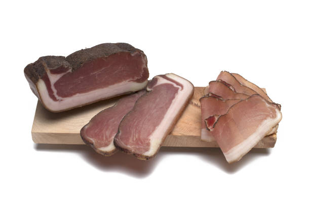 Close up of a large piece of South Tyrolean square bacon next to thicker sheaves and thin leaves on a wooden cutting board against a white background
