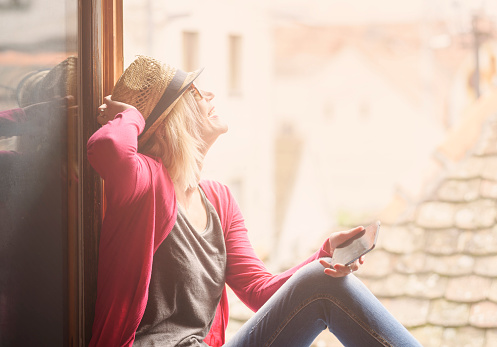 Young happy hipster woman in casual fashion clothing and summer stylish hat sitting at the home window, holding smartphone, laughing and enjoy the sun. Youth lifestyle concept. Retro style filter.