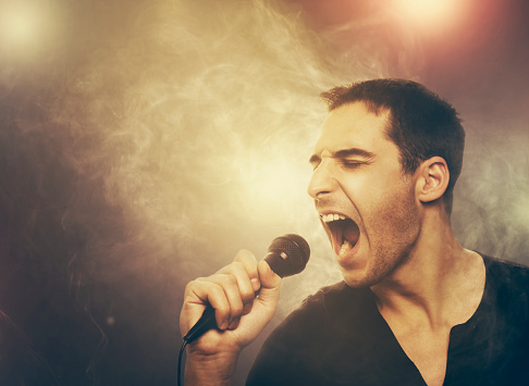 Young man singer holding microphone and singing karaoke music in night club. Rock star screaming on the concert with smoke and stage lights.