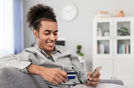 Young multiracial man with afro hair holding a mobile phone and credit card and making online shopping through mobile app, while sitting on the sofa in his living room.
