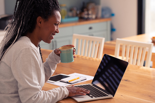 Freelancer woman typing on a laptop computer, sitting at the kitchen table, drinking coffee.