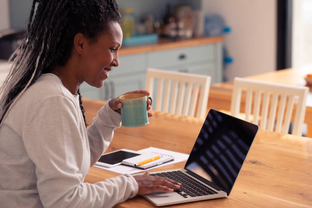 Freelancer woman typing on a laptop computer, sitting at the kitchen table, drinking coffee. A woman in casual clothing using a laptop and smiling while comfortably sitting at the kitchen table, drinking coffee. Freelance Writing Jobs stock pictures, royalty-free photos & images