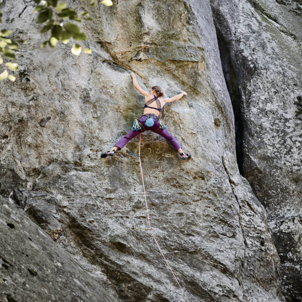Young woman climbing difficult route on a high rock with rope. Fearless climber training rock climbing on summer day. Concept of extreme sport, adventures and active lifestyle.