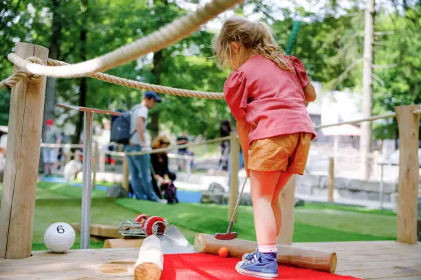 Cute preschool girl playing mini golf with family. Happy toddler child having fun with outdoor activity. Summer sport for children and adults, outdoors. Family vacations or resort