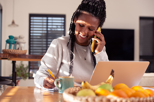 A businesswoman woman calling on a smartphone and writing to notepad, using a laptop, sitting in the kitchen. Female working from distance at home. Online business career.