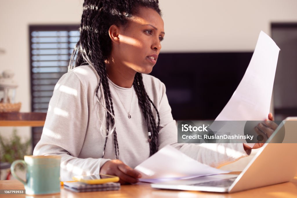 Female entrepreneur working at home. The woman reading documents. African-American woman sitting at the desk with laptop, reading, reviewing few documents. Female working from distance at the home office. Online business career. Law Stock Photo