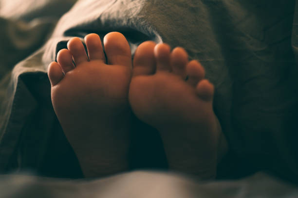 Bare children's feet stick out from under the blanket. Warm and cozy photo in a dark key. To wallow and rest in bed on a cloudy day. The child plays in bed.