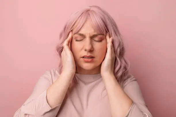 Young woman feeling stressed over pink background. Suffering migraine.