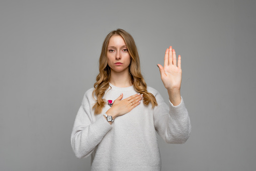 Portrait of very responsible and honest woman giving promise, making solemn vow in ceremonial tradition with raised hand, wears white sweater, gray background