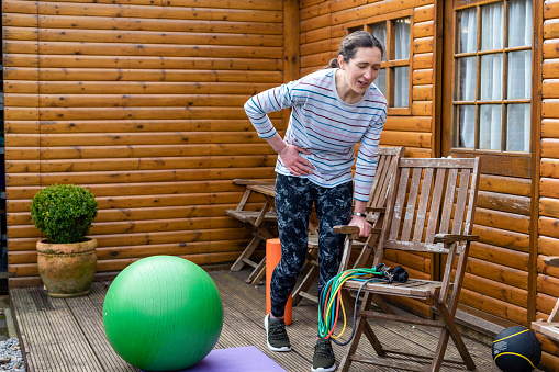 Woman standing in her garden in the North East of England exercising. She is bending over holding her side with a pained expression.