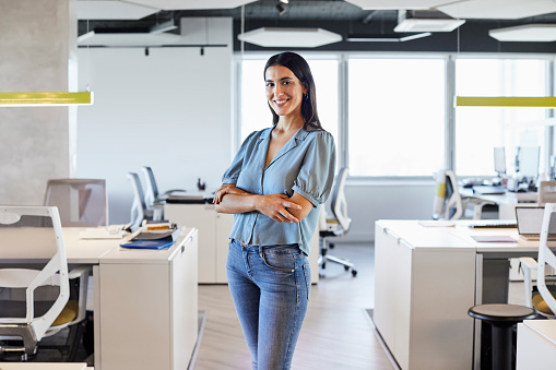 Portrait of smiling businesswoman in office. Female entrepreneur with arms crossed is standing at workplace. She is wearing smart casuals.