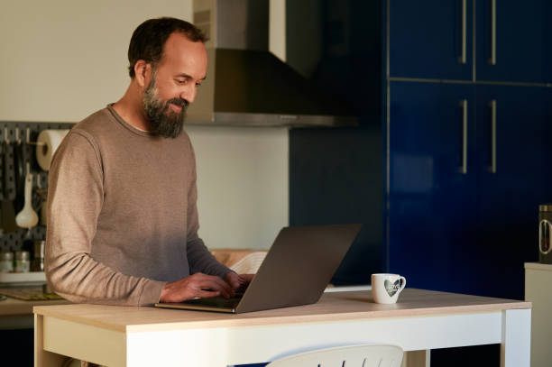 istock smiling middle-aged bearded man using his laptop in the kitchen of his home while drinking coffee 1367813288