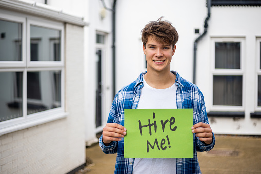 A young man, looking for employment and holding up a sign with 'hire me!' on it. He is looking at the camera with a smiley expression on his face.