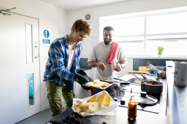 Making Garlic Bread! A side view shot of male uni student friends standing in their kitchen, they are wearing casual clothing and preparing a meal together. One man is carrying a tray of garlic bread out of the oven wearing oven mitts. middlesbrough stock pictures, royalty-free photos & images