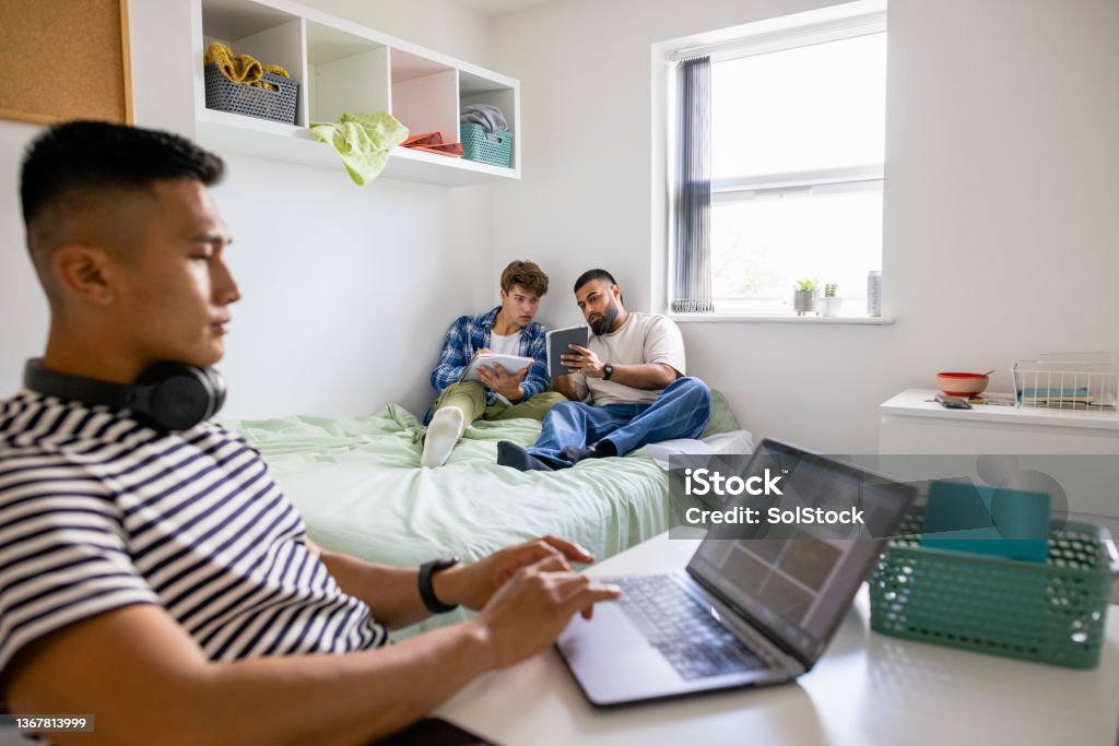Uni Students Studying Together A small group of male uni student friends sitting in their dorm bedroom are studying together while sitting on the bed, one man is sitting at his desk. Studying Stock Photo