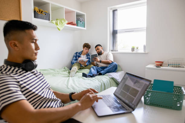 Uni Students Studying Together A small group of male uni student friends sitting in their dorm bedroom are studying together while sitting on the bed, one man is sitting at his desk. middlesbrough stock pictures, royalty-free photos & images