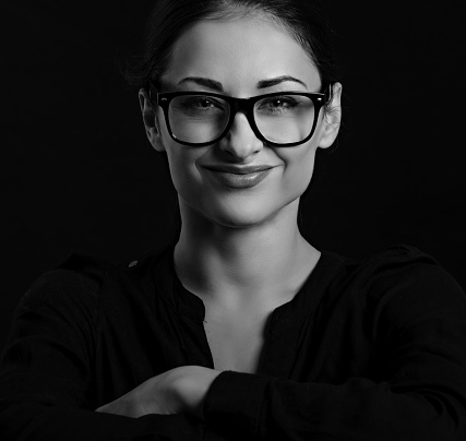 Beautiful thinking smiling happy business woman  looking happy in eye glasses and holding spectacles in casual black t-shirt on black background. Closeup portrait. Black and white