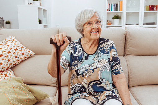 Portrait of happy mature woman in eyeglasses holding cane while sitting on sofa at home. Cheerful smiling aged grandmother looking at camera. Lifestyle concept.