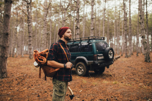 istock Millennial man hipster with beard wearing backpack, holding axe and firewood in forest with car on background. 1367823791
