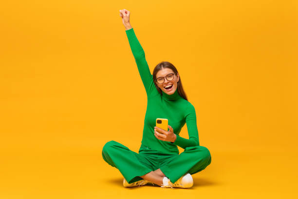 Winner. Excited young woman sitting on floor with her phone, shouting yes, isolated on yellow Winner. Excited young woman sitting on floor with her phone, raising one hand in the air saying yes, isolated on yellow background excited woman yellow stock pictures, royalty-free photos & images
