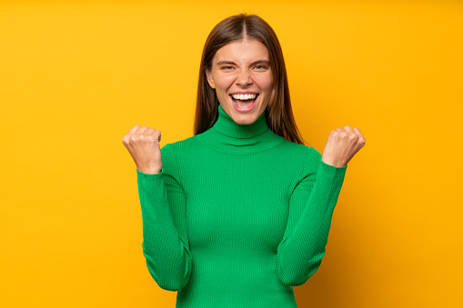 Yes! Young happy celebrating woman is a winner, isolated on yellow background