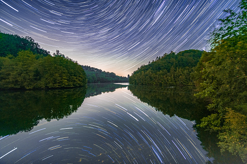 Startrails of night sky is reflecting in the perfectly still lake Kleine Dhünn Dhünntalsperre surrounded by green forest landscape in Bergisches Land region in western germany